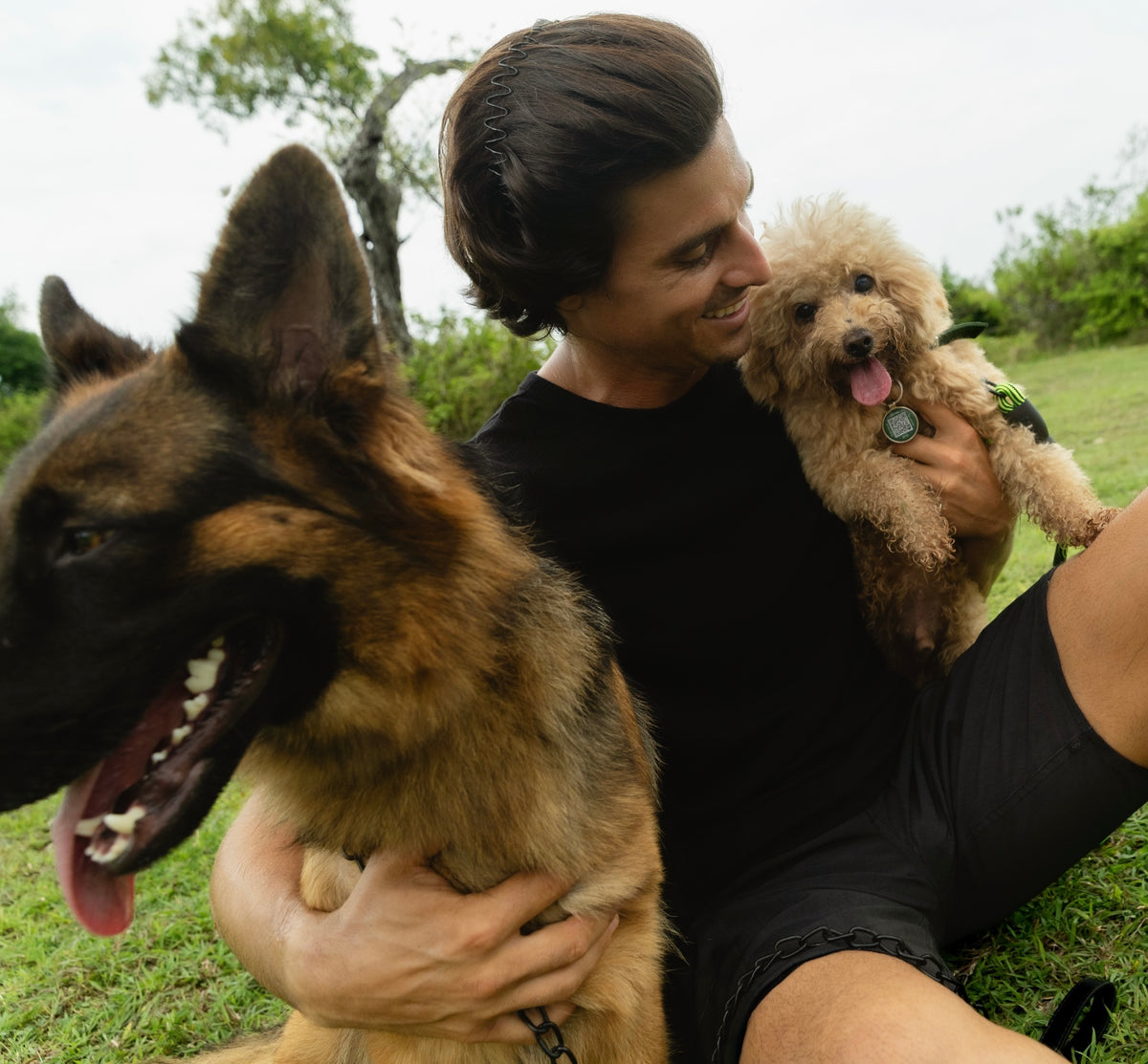 Man smiling while holding a small poodle wearing a Supernormal tag, with a German Shepherd beside him
